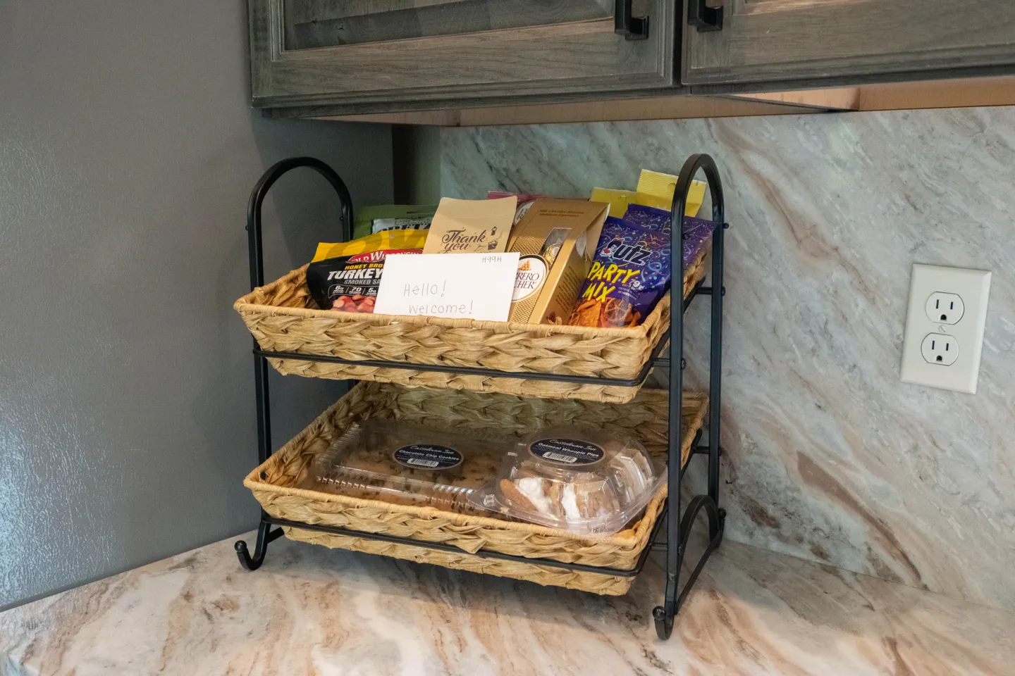 Two-tier woven basket filled with welcome snacks and a greeting card on a kitchen counter.