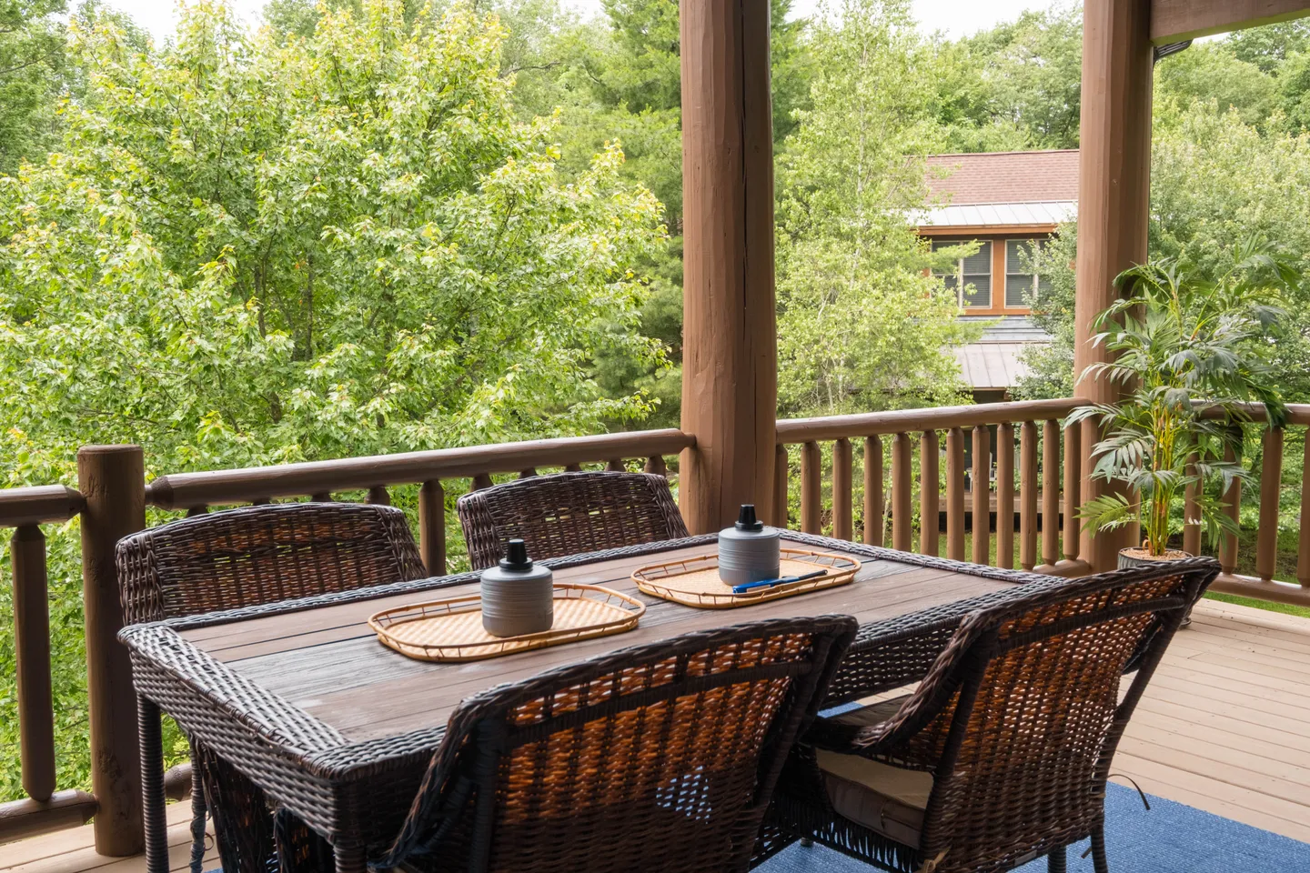 Outdoor covered deck featuring a wicker dining table and chairs, overlooking lush green trees.