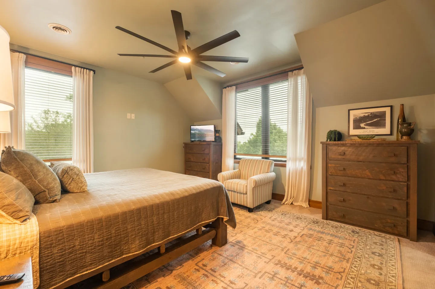 Bedroom with a wooden bed, patterned area rug, striped armchair, wooden dressers, and a large ceiling fan.