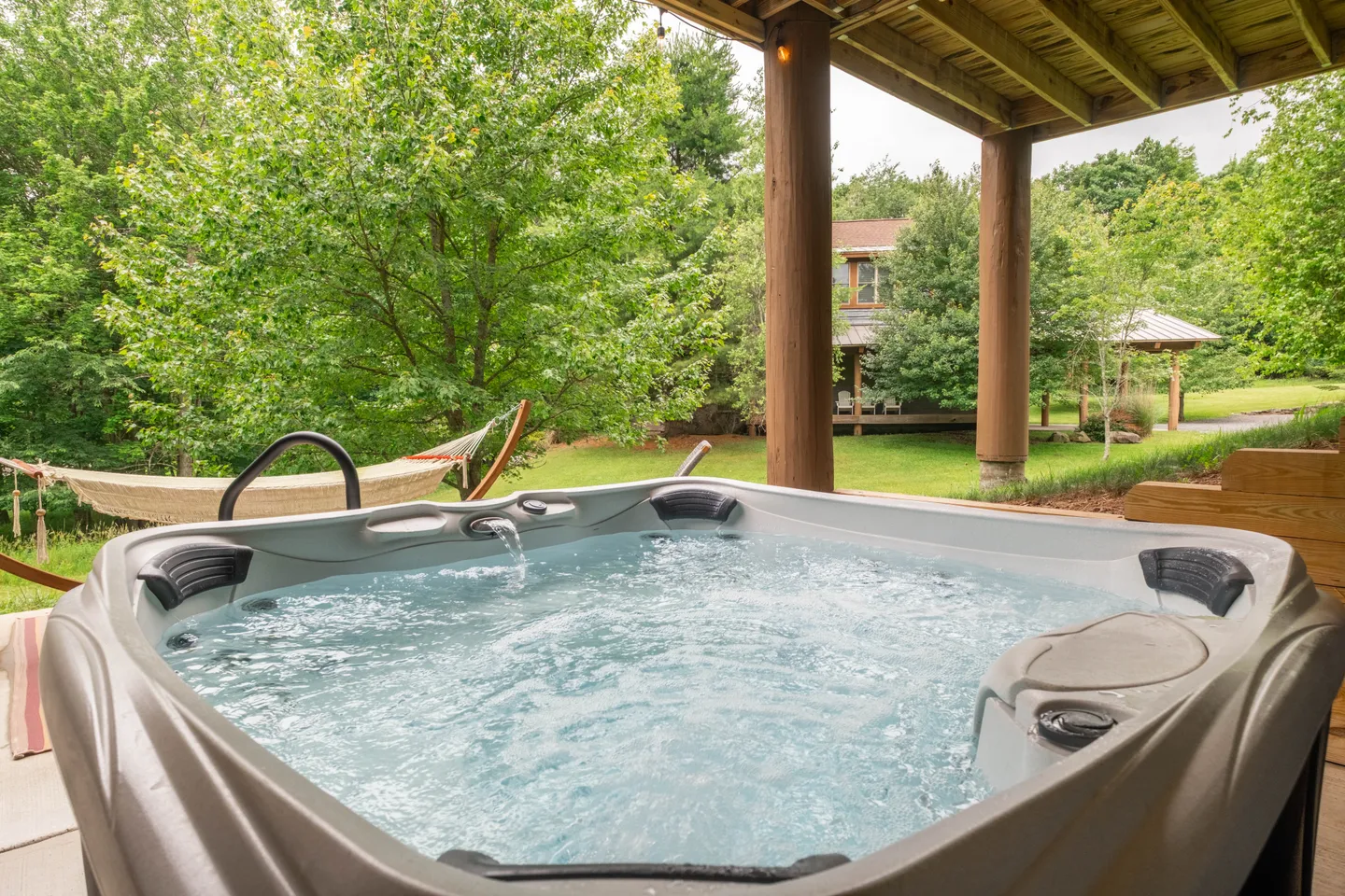 Outdoor hot tub filled with bubbling water on a covered patio overlooking a grassy lawn with trees and a hammock.
