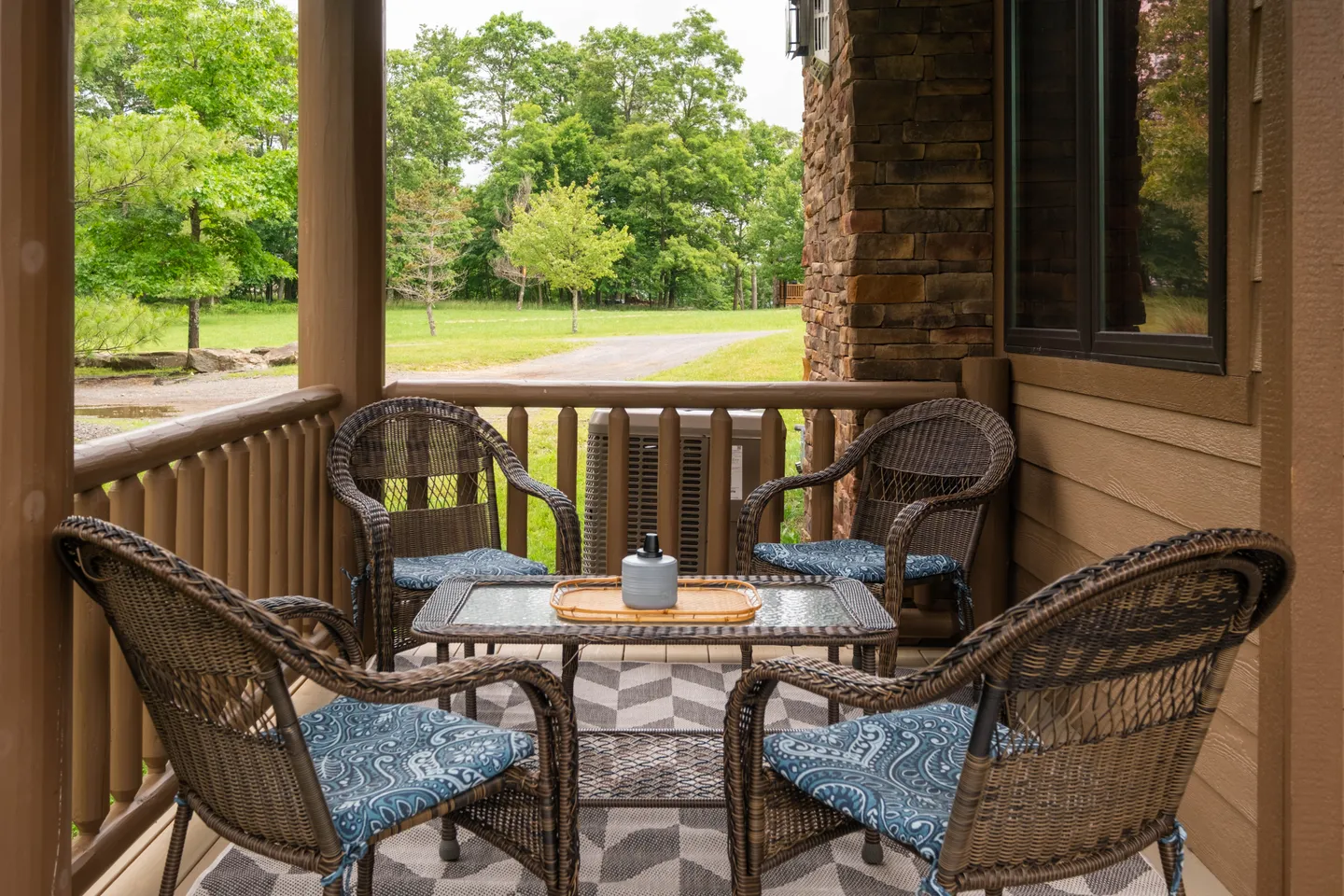 Covered porch with four wicker armchairs, a glass-topped coffee table, and an outdoor rug looking out onto trees and a gravel driveway.