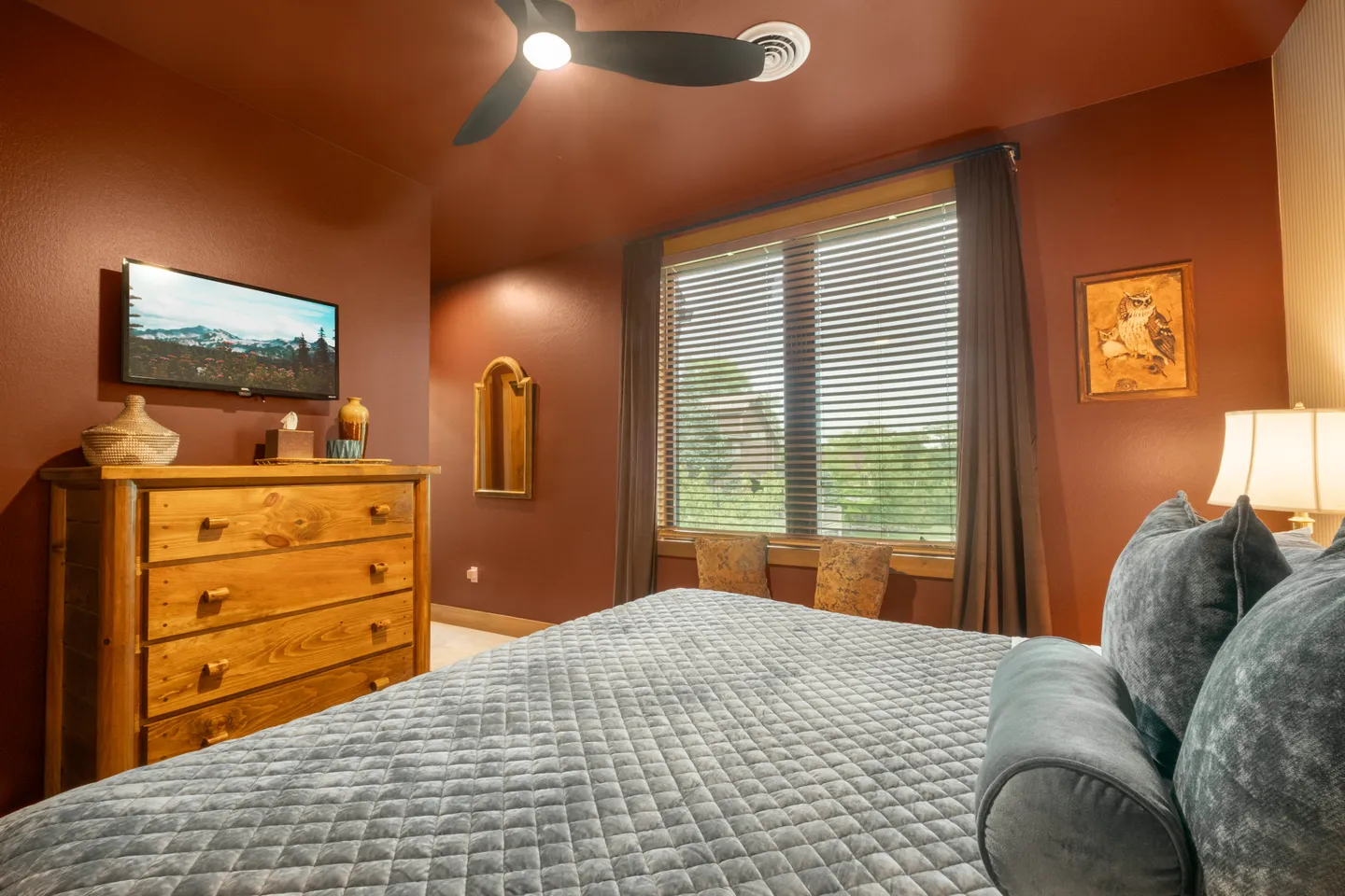 Bedroom with rust-colored walls, a wooden dresser, a mounted TV, a window with blinds, and a bed with a quilted grey coverlet.