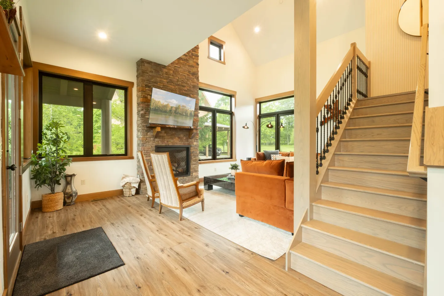 Open living area with wood-look floors, a stone fireplace, an orange sofa, armchairs, and a light wood staircase.