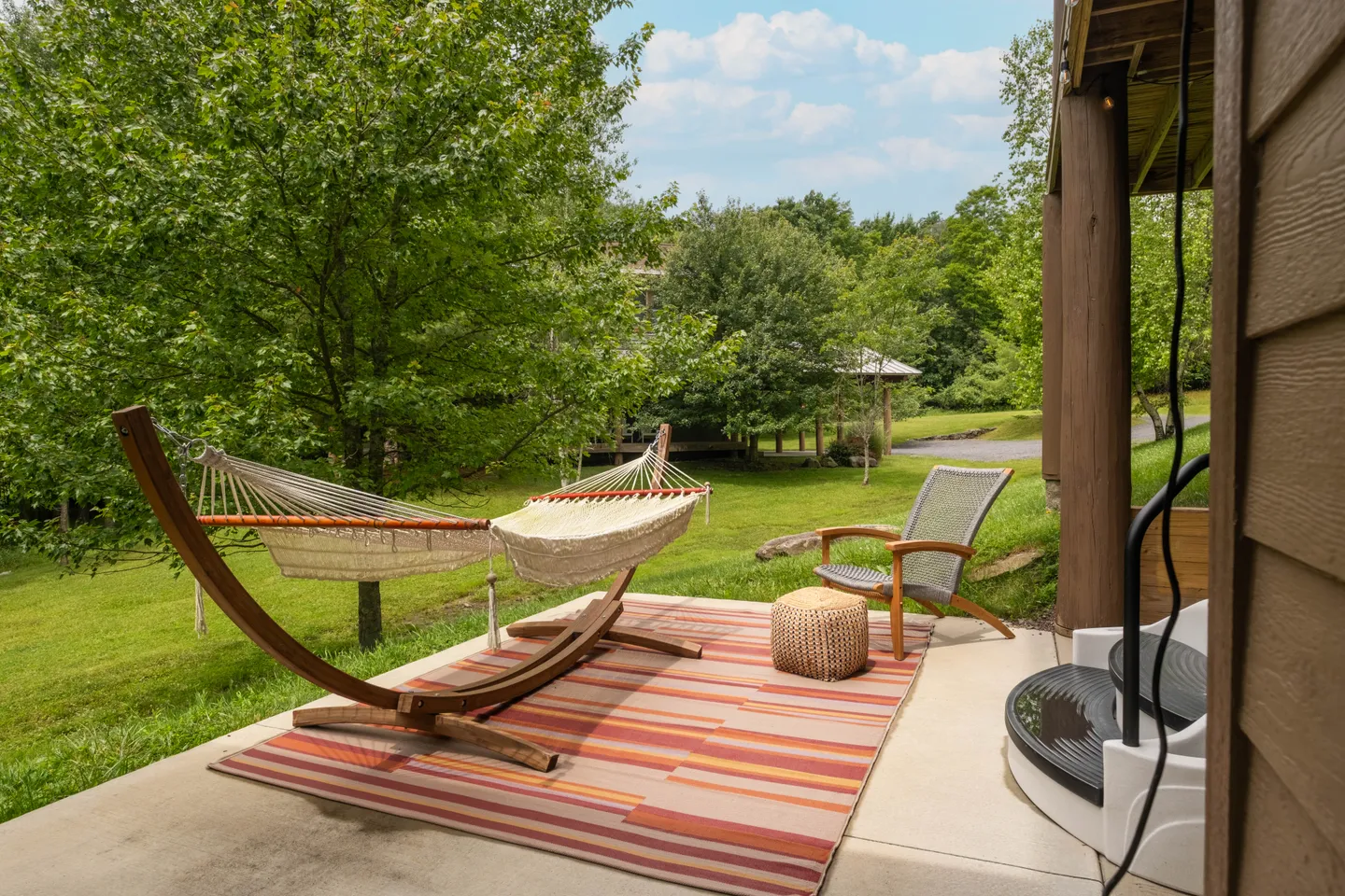 Outdoor patio with a wooden-frame hammock, an armchair, an ottoman, and a striped area rug overlooking a grassy lawn and trees.