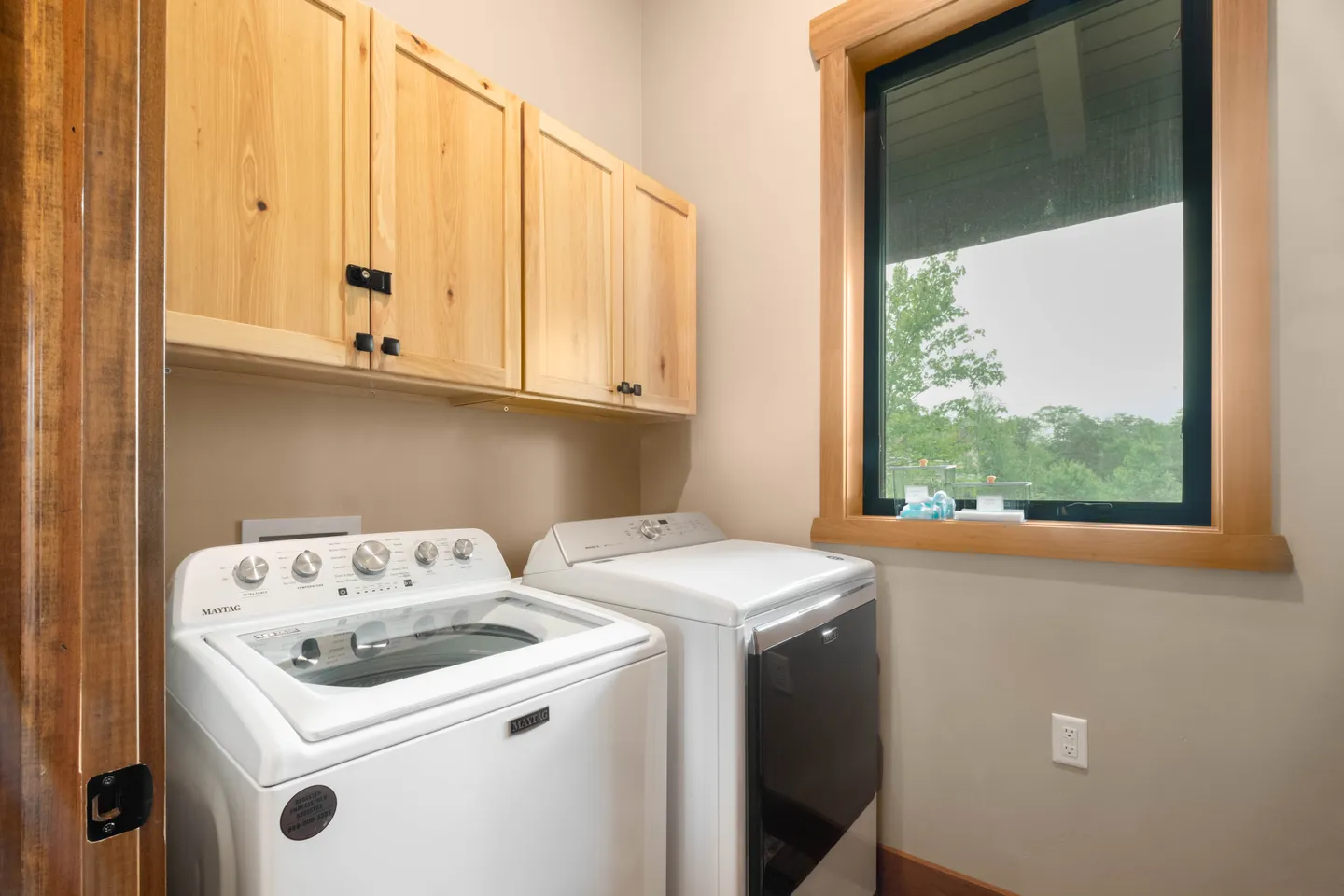 A laundry room with a white top-loading washing machine, a white dryer, light wood upper cabinets, and a window trimmed in wood.