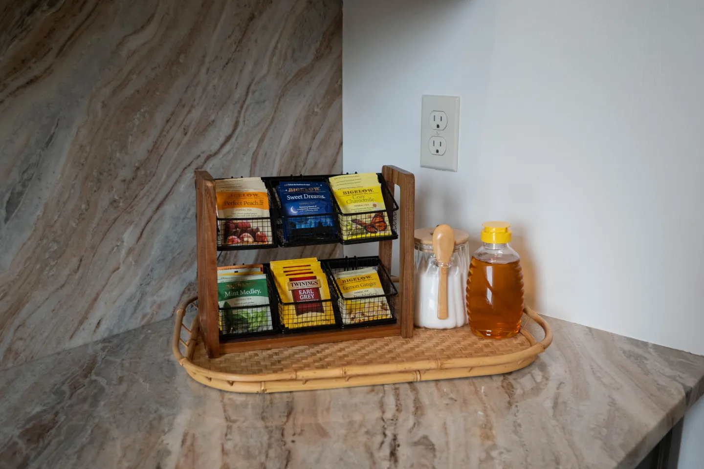 Close-up of a tea station on a countertop featuring an assortment of packaged tea bags in a tiered wooden caddy alongside sugar and honey.