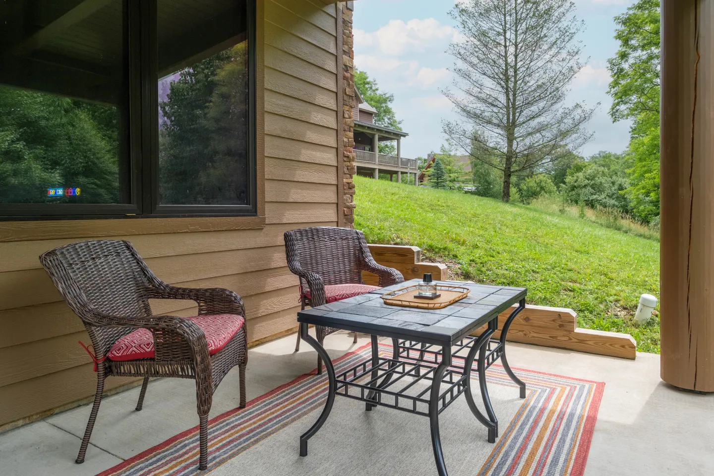 A concrete patio with two wicker chairs, a small coffee table, and an outdoor rug overlooking a grassy hillside.