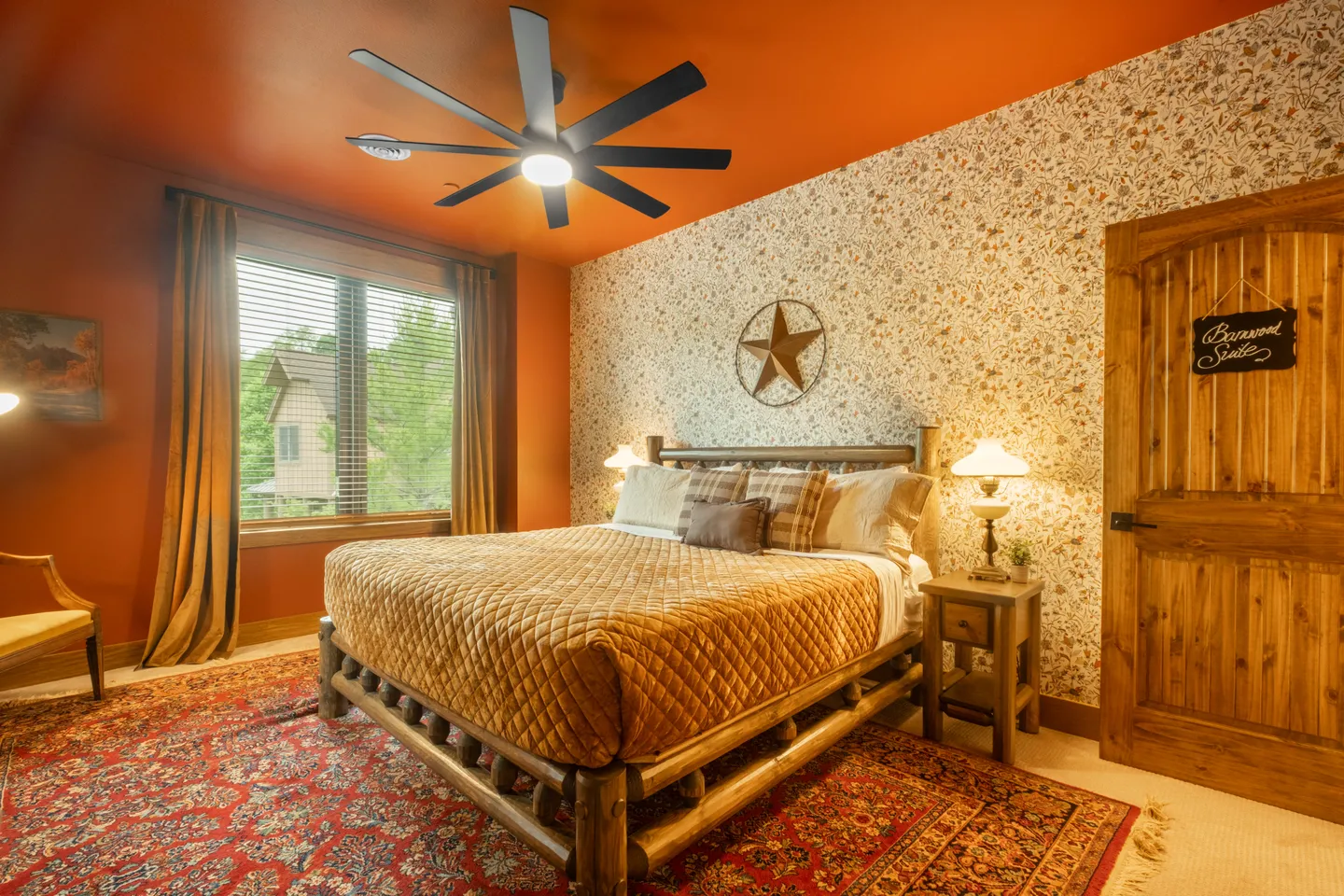 Bedroom featuring a log-style bed with quilted bedding, a ceiling fan, floral wallpaper on an accent wall, and burnt orange paint.
