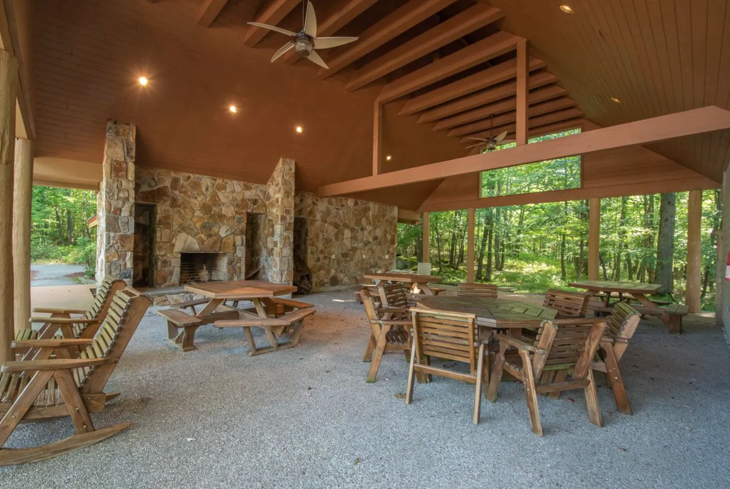 Open-air wooden pavilion featuring a stone fireplace, wood picnic tables, rocking chairs, and views of surrounding green woods.