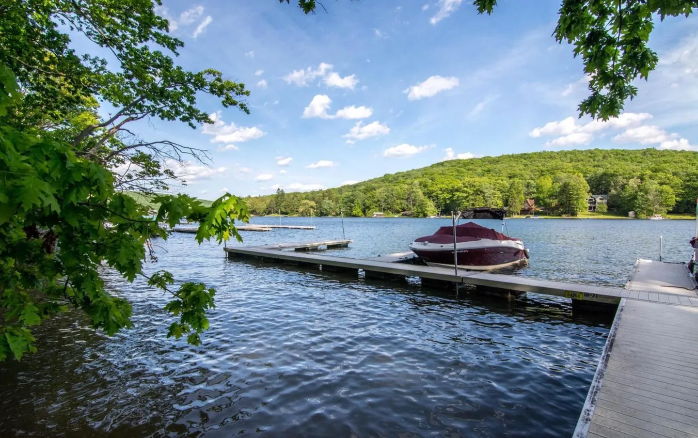 A wooden boat dock extending into a wide lake with a covered boat moored beside it, surrounded by lush green hills.