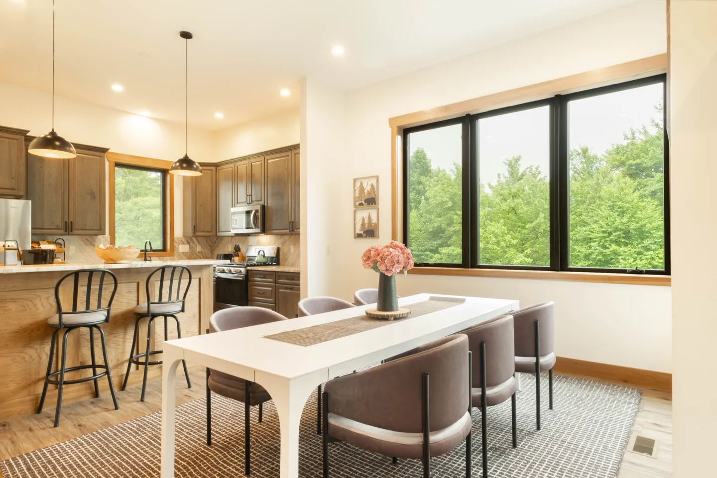 Dining area with a long white table and upholstered chairs adjacent to an open kitchen with bar seating and large windows overlooking trees.