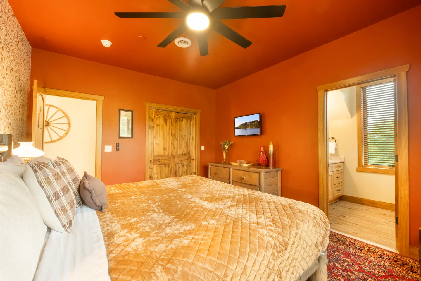 Bedroom with bold terracotta walls, ceiling fan, large bed with golden quilted bedding, wooden dresser, wall-mounted TV, and an open doorway to an en-suite bathroom.