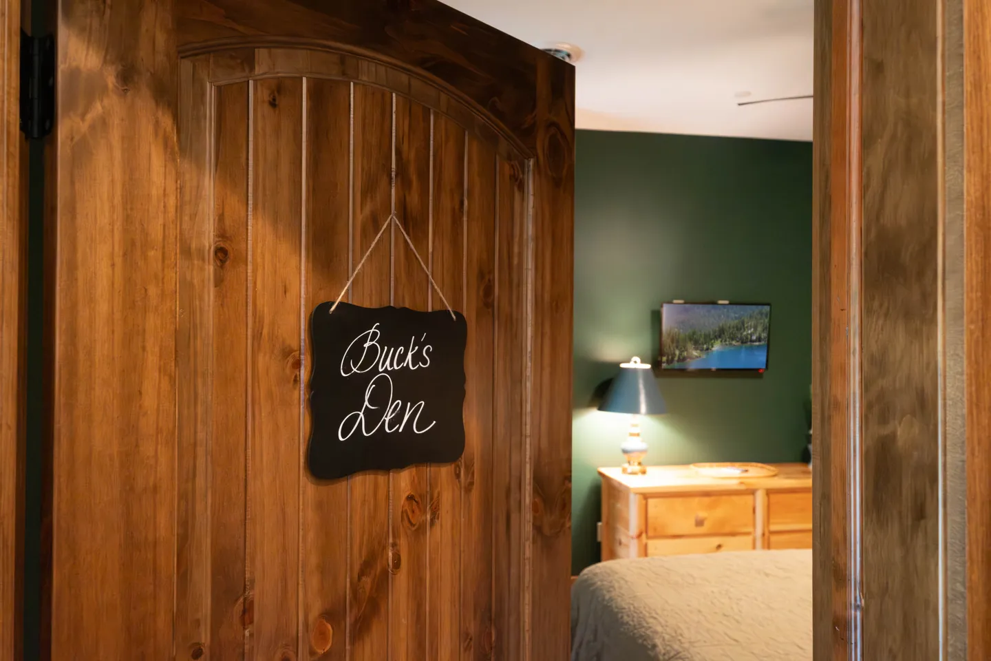 Open wooden paneled door with a chalkboard sign reading Buck's Den looking into a bedroom with green walls.