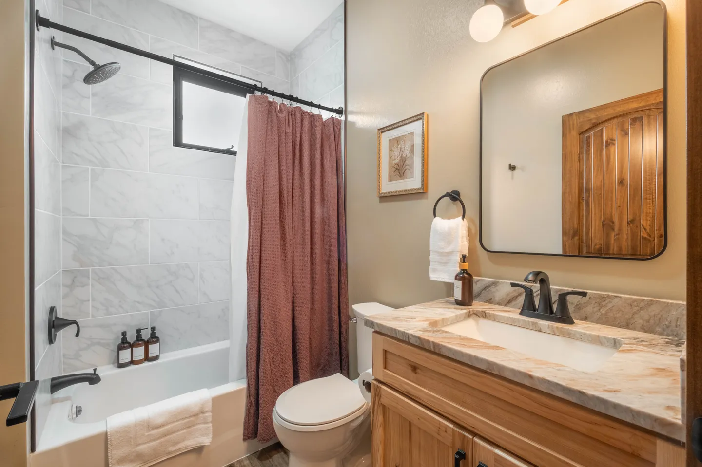 Bathroom with a wood vanity, marble-style countertop, toilet, and tiled shower-tub combination with a dark curtain.
