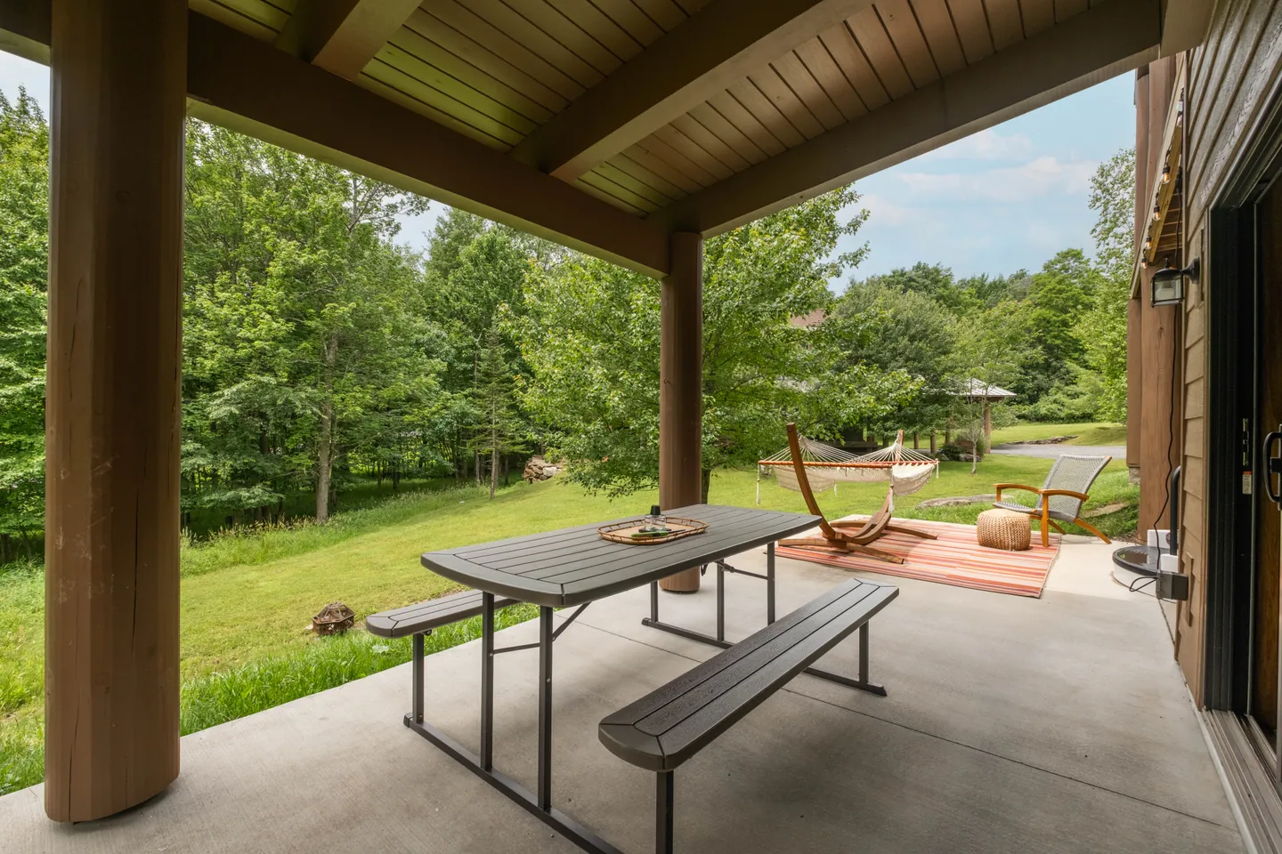 Covered concrete patio with a picnic table and benches overlooking a grassy yard, trees, and a hammock.