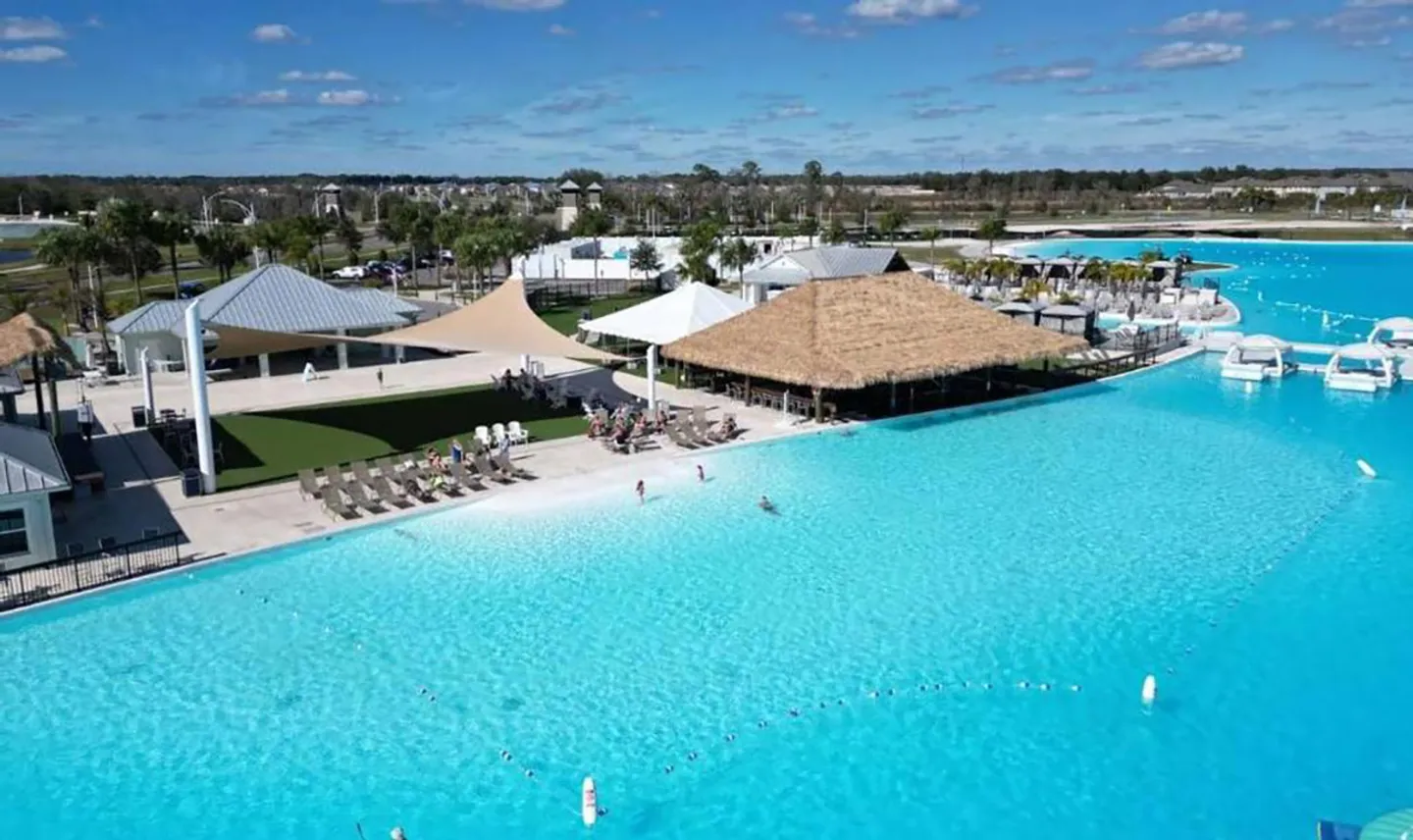 Aerial view of a large turquoise lagoon pool featuring a sandy shoreline, lounge chairs, a thatched-roof pavilion, and community amenity buildings under a blue sky.