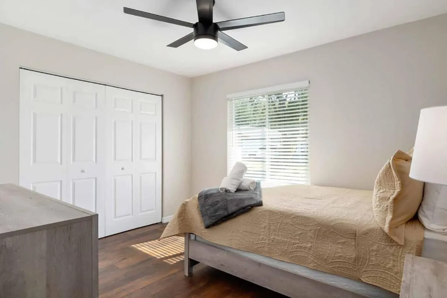 Bedroom featuring a made bed with a tan quilt, a wooden dresser, a window with horizontal blinds, and a white bifold closet door.