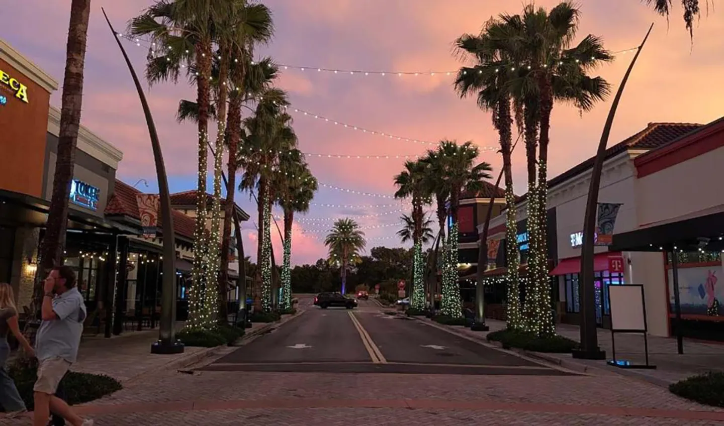 Street view at dusk lined with palm trees wrapped in string lights and buildings on both sides under a pink sky.