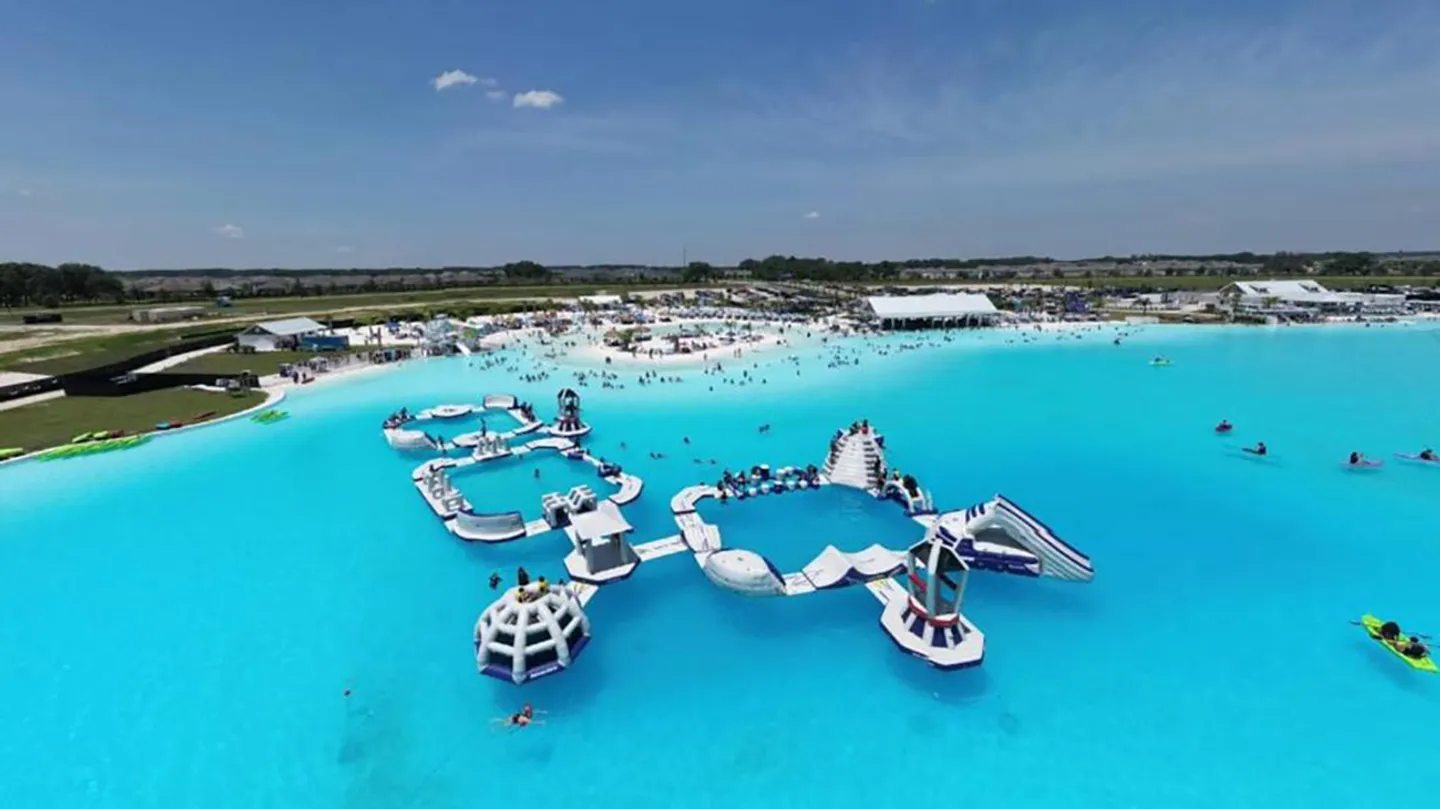 Aerial view of a large turquoise lagoon waterpark with inflatable obstacle courses, paddleboarders, and sandy beach areas.