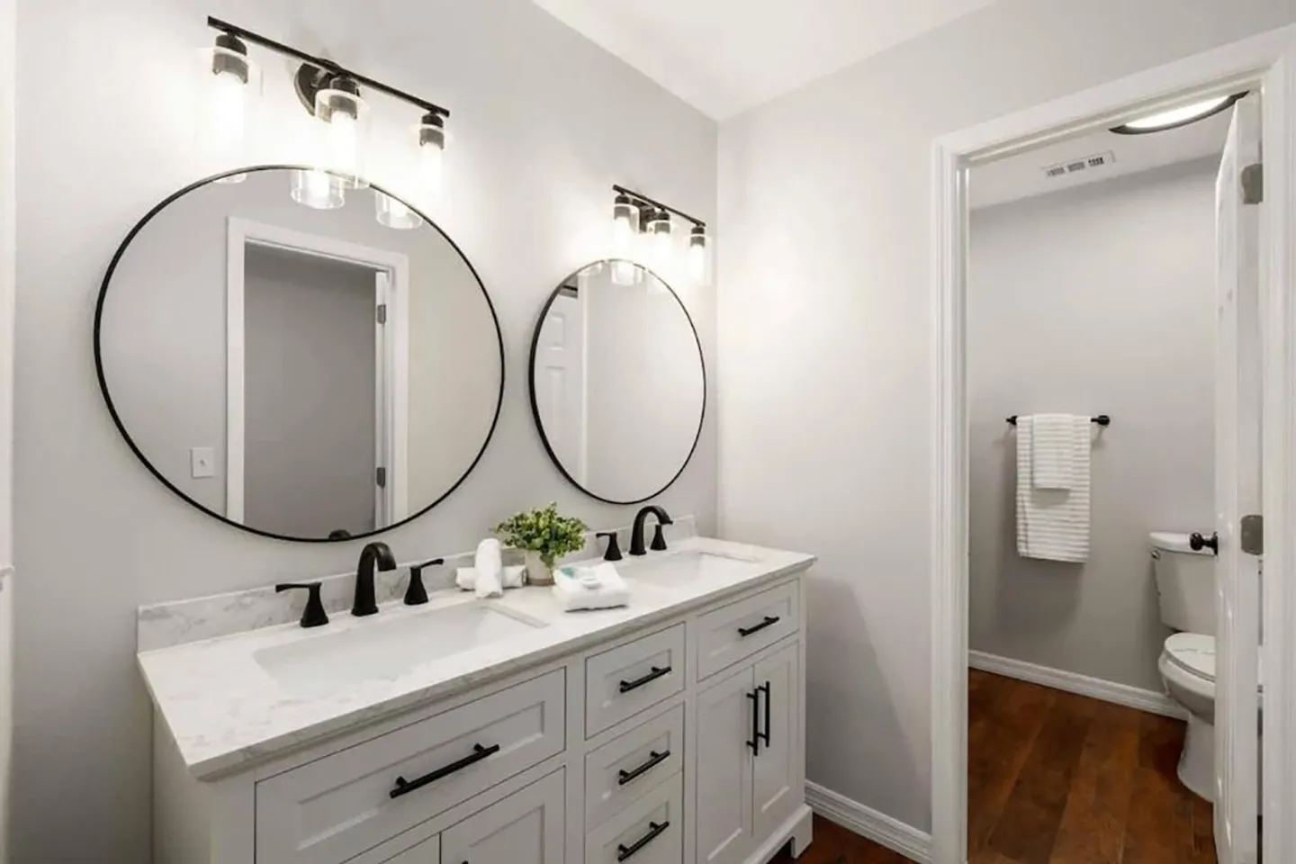 Bathroom vanity with double sinks, two round mirrors, black fixtures, and a view into an adjacent toilet room.