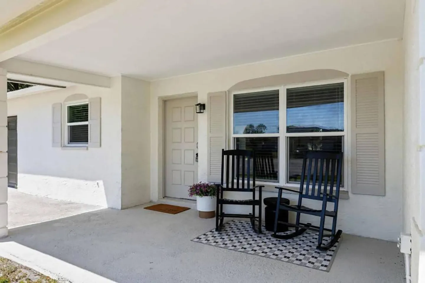 Covered front porch with two black rocking chairs on a patterned rug in front of shuttered windows.