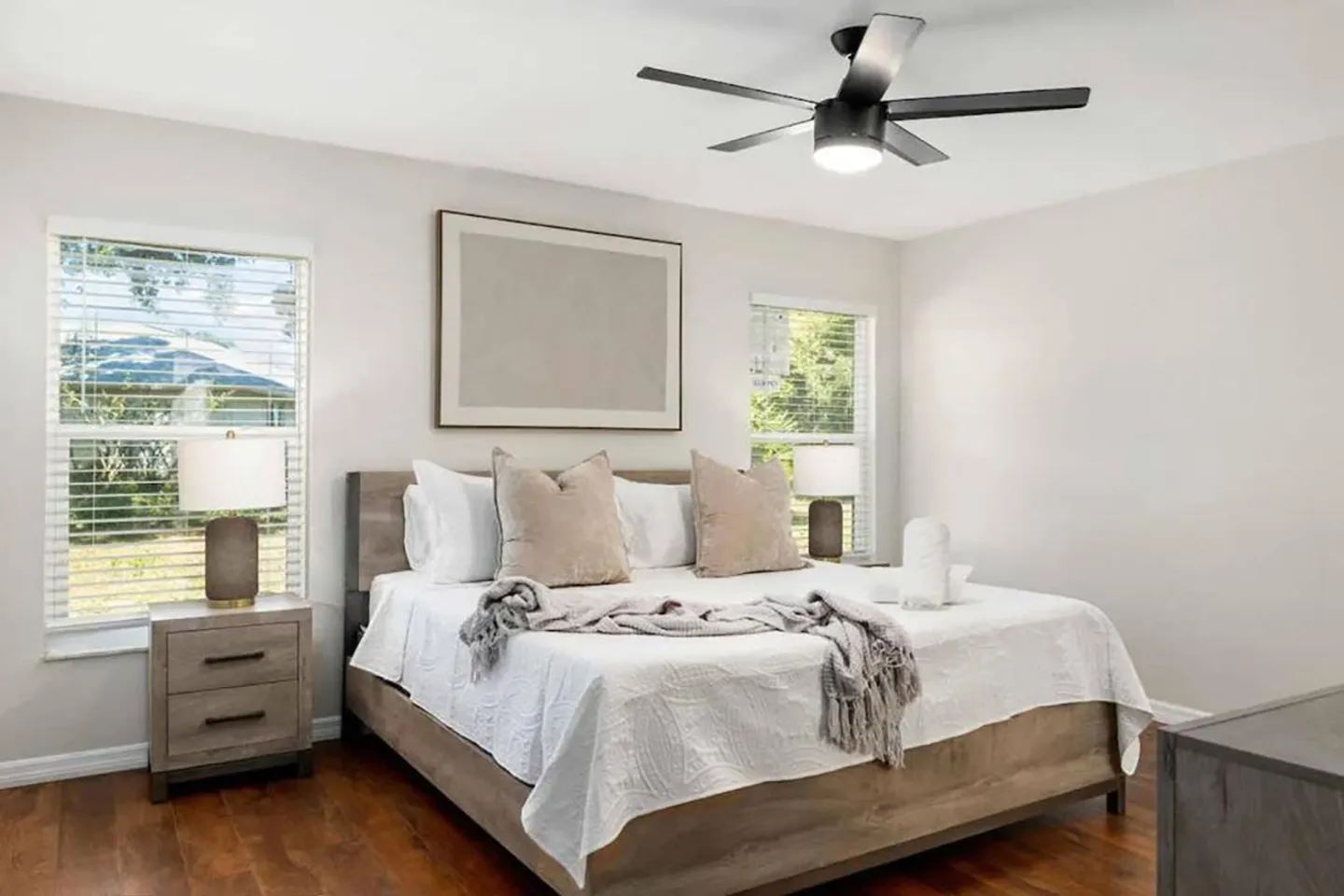 Bedroom with a wooden bed frame, light bedding, two windows, nightstands with lamps, and a modern ceiling fan.