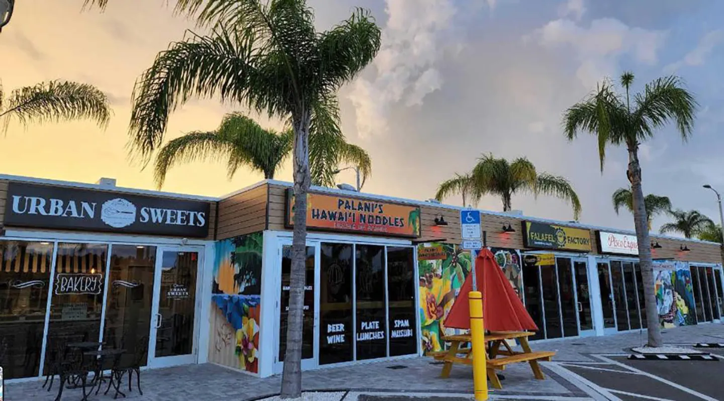 Exterior of an outdoor strip mall featuring storefronts such as Urban Sweets and Palani's Hawai'i Noodles under palm trees at dusk.