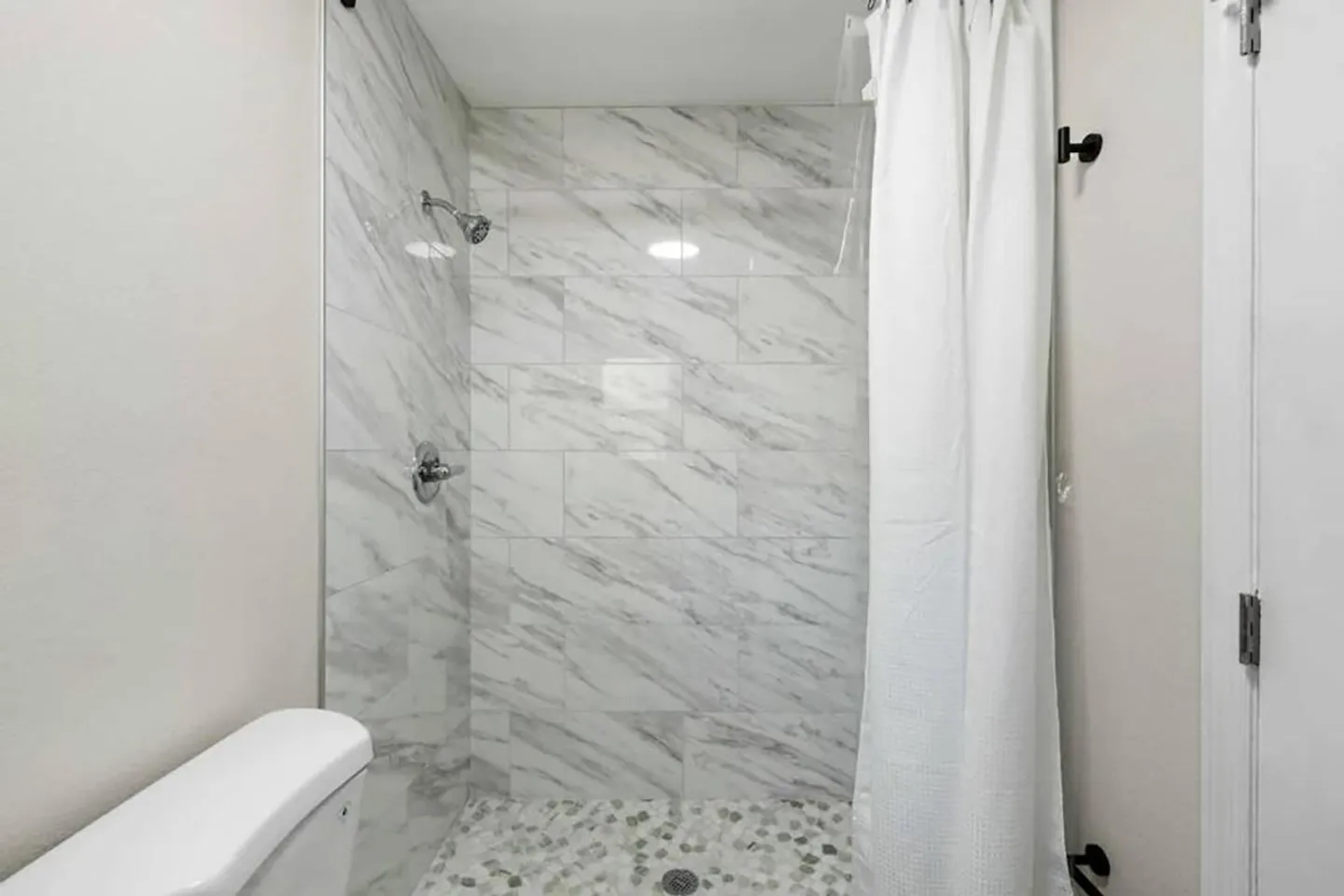 Walk-in shower with marble-look wall tiles, a pebble floor, and a white curtain next to a toilet tank.
