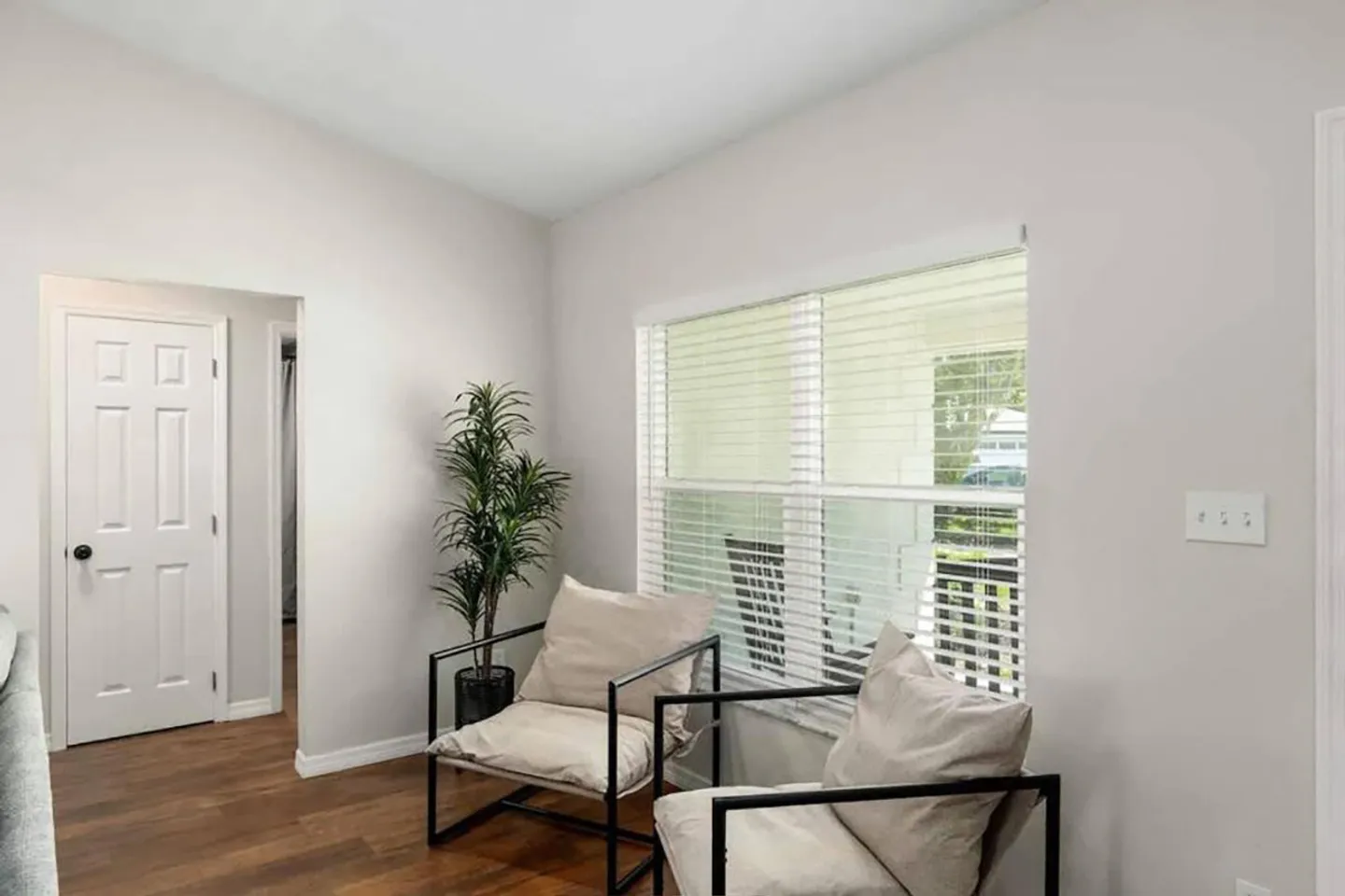 Seating area with two modern accent chairs near a window with blinds, an indoor potted plant, and wood-look flooring.