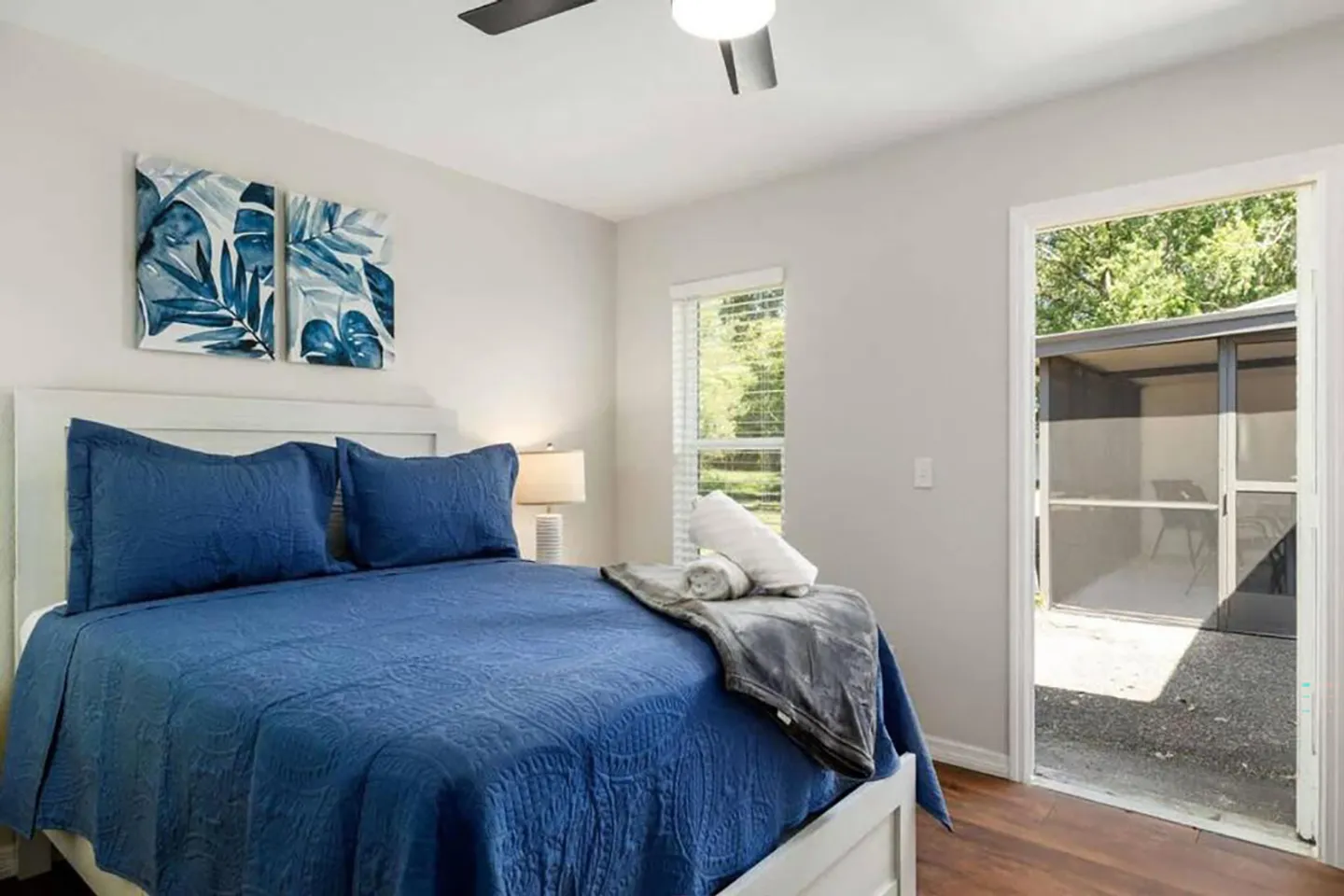 Bedroom with a blue-quilted bed, a ceiling fan, a window, and an open doorway leading outdoors to a screened structure.