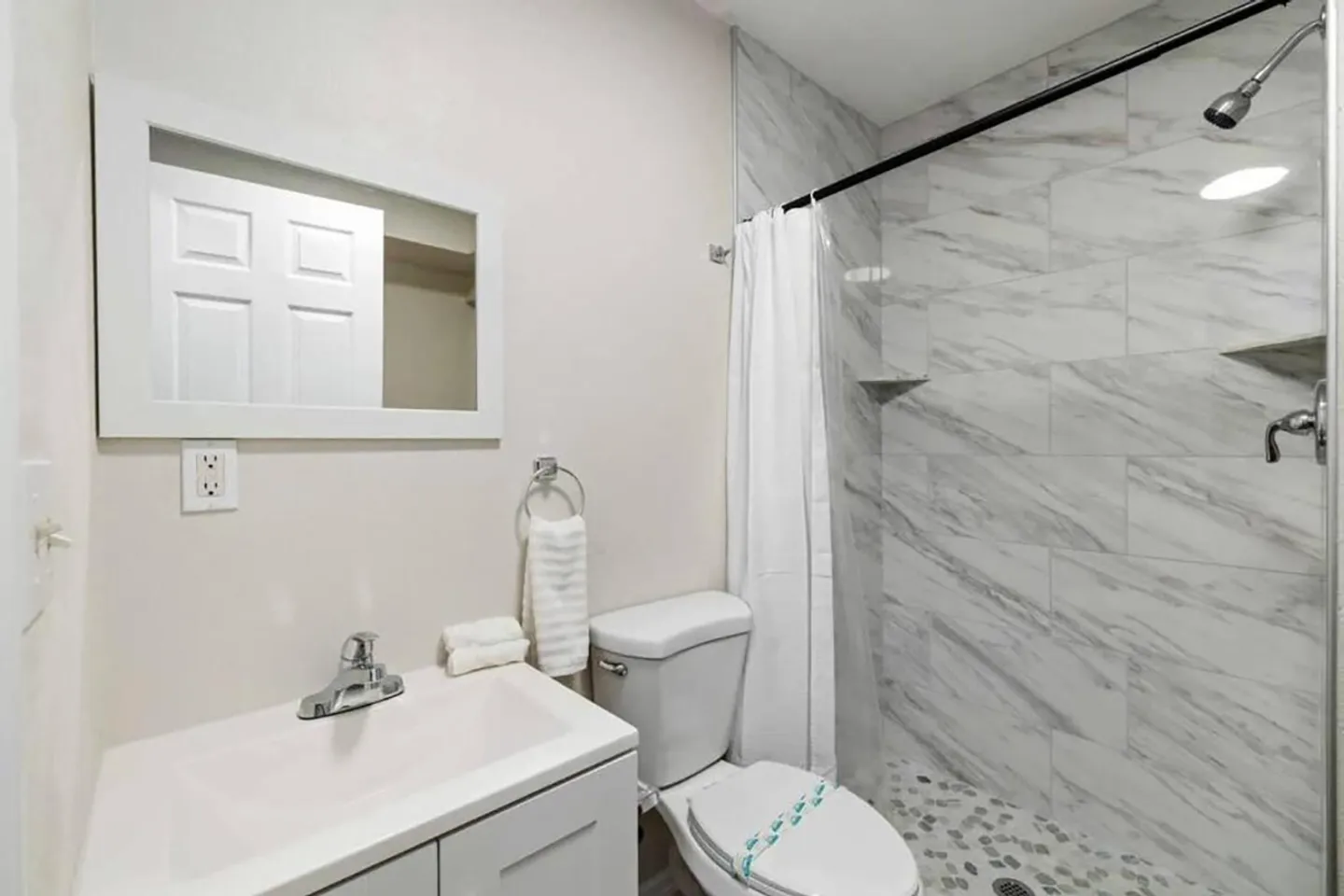 Bathroom with a white vanity, framed mirror, toilet, and walk-in shower lined with marble-patterned tiles.