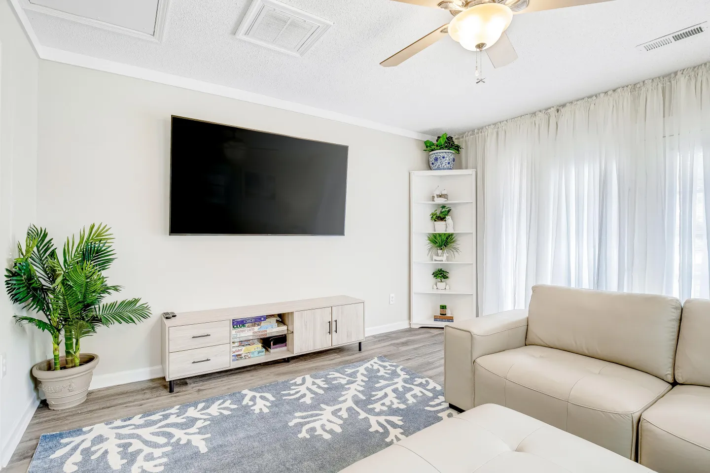 Bright living room with a wall-mounted TV over a low credenza, corner shelving, patterned blue area rug, and cream-colored leather seating.