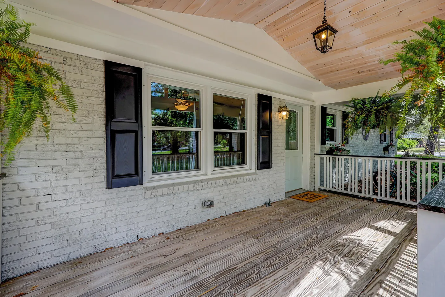 Front porch of a house with wooden plank flooring, painted brick siding, black shutters, and hanging fern plants.