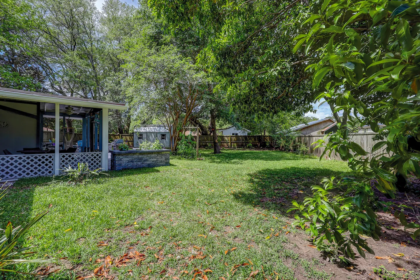 Fenced grassy backyard featuring mature shade trees, a storage shed, and an adjacent covered patio structure.