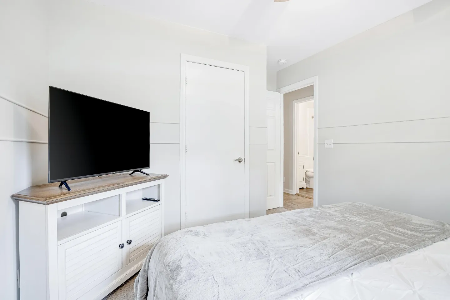 Bedroom featuring a bed with a light-colored throw blanket, a white media console with a flat-screen television, and a doorway opening to a bathroom.