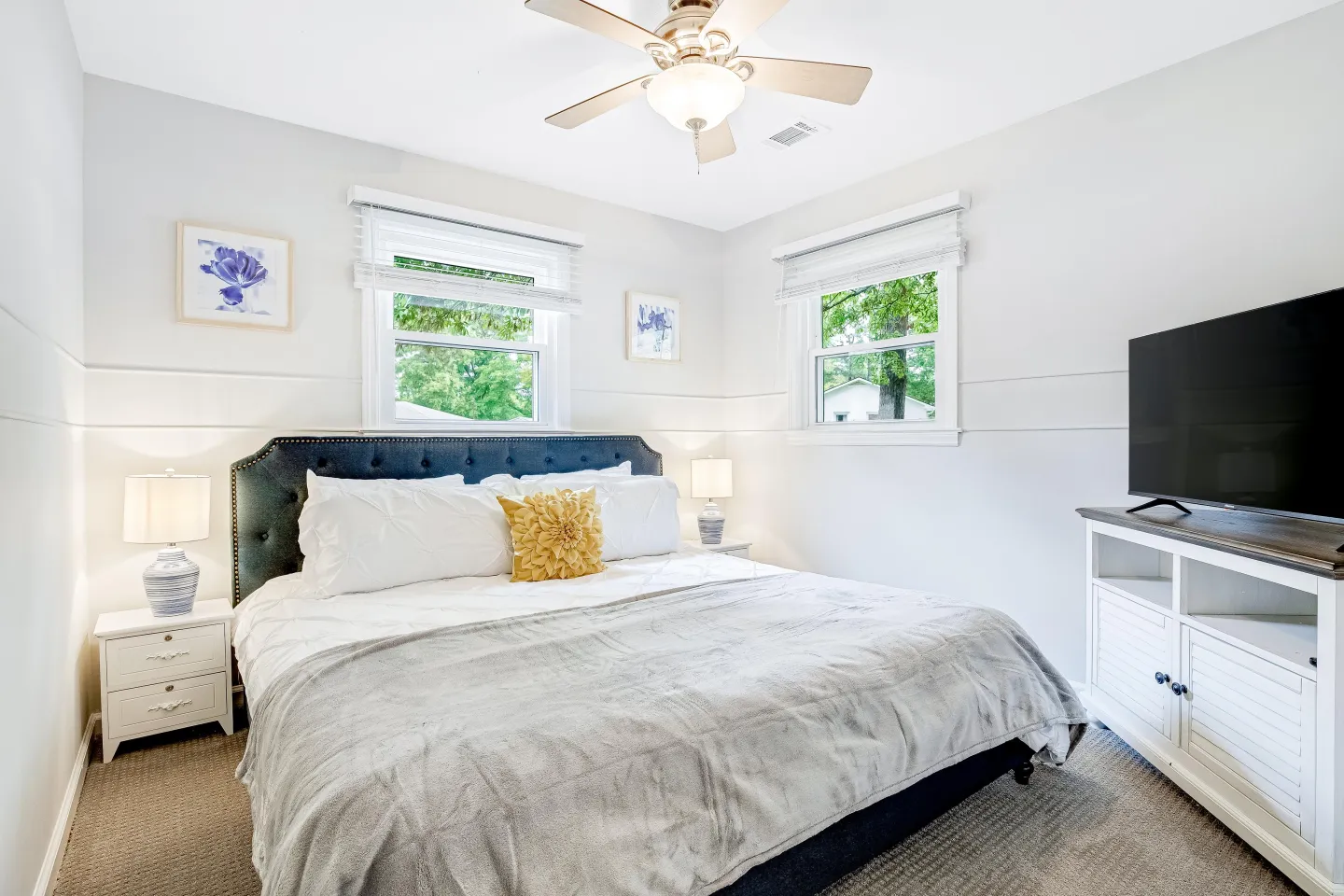 Bedroom with a tufted headboard on the bed, two windows, nightstands with lamps, a ceiling fan, and a TV on a console.