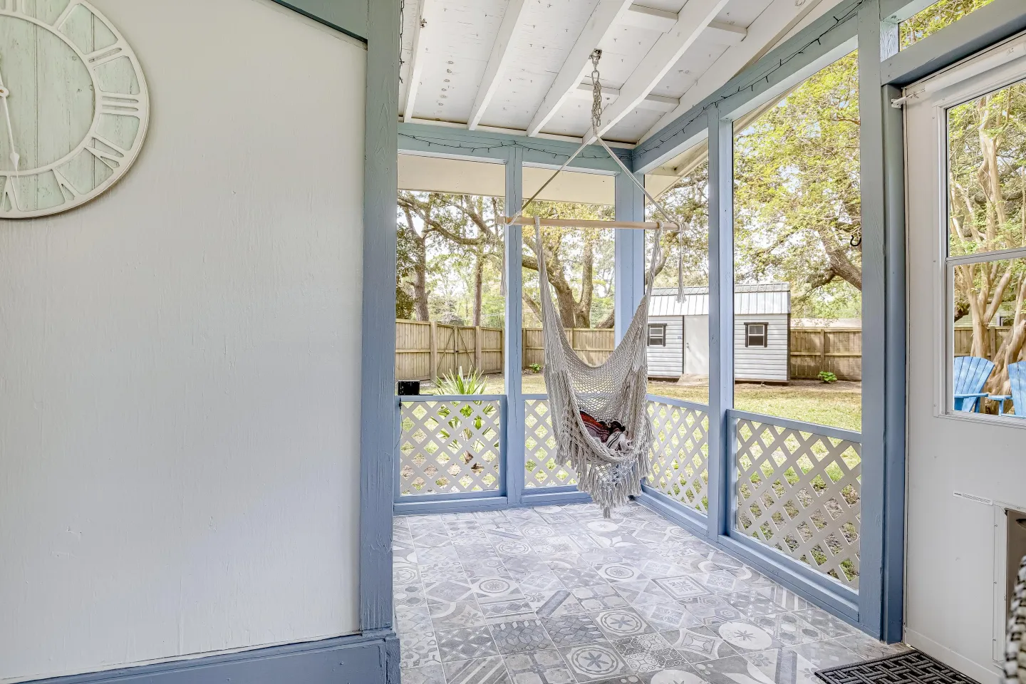 Screened porch with blue-painted frame, patterned tiled floor, and a hanging macrame hammock chair overlooking a grassy fenced yard.