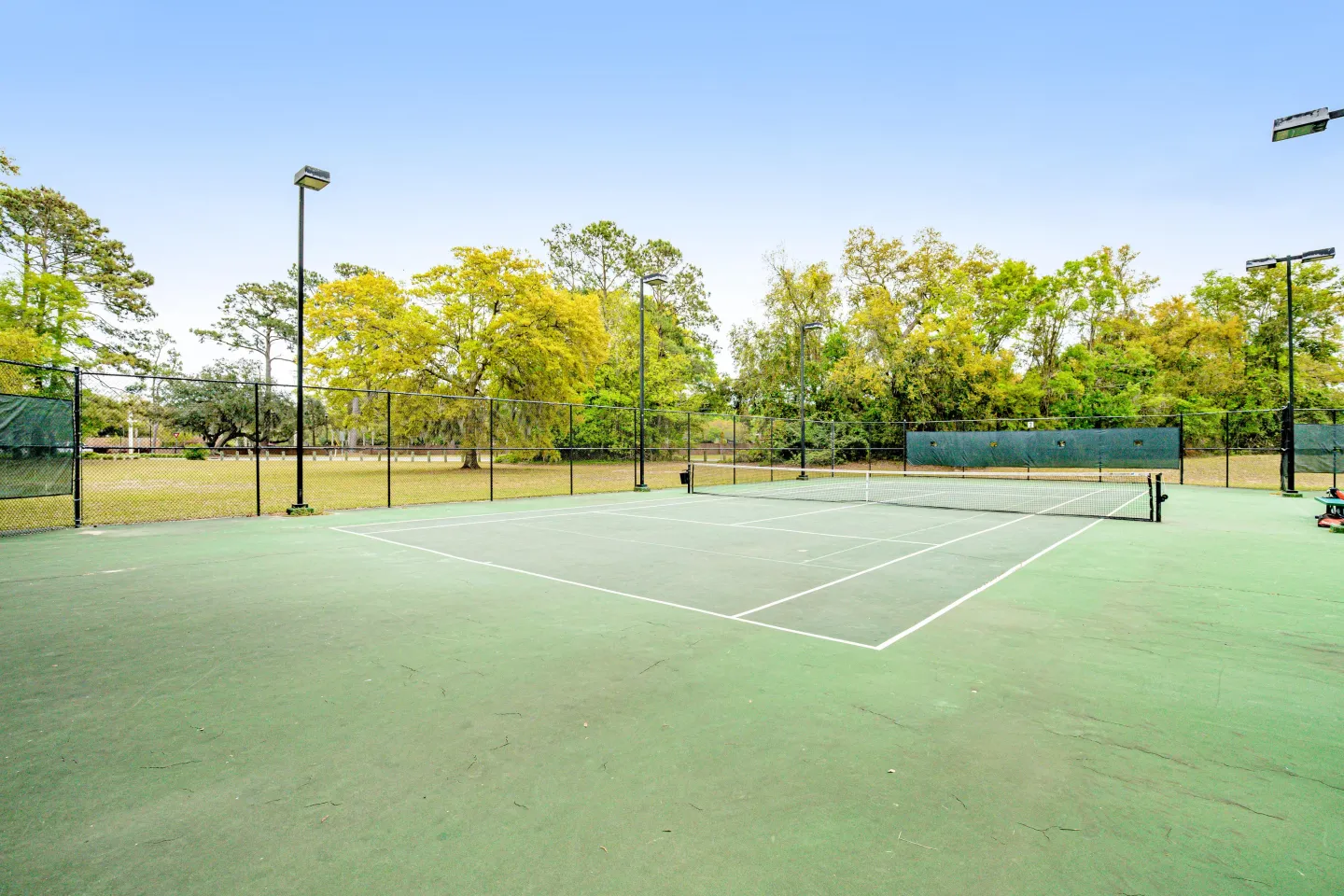 Outdoor tennis court enclosed by chain-link fencing with light poles and surrounded by green and yellow trees under a clear sky.