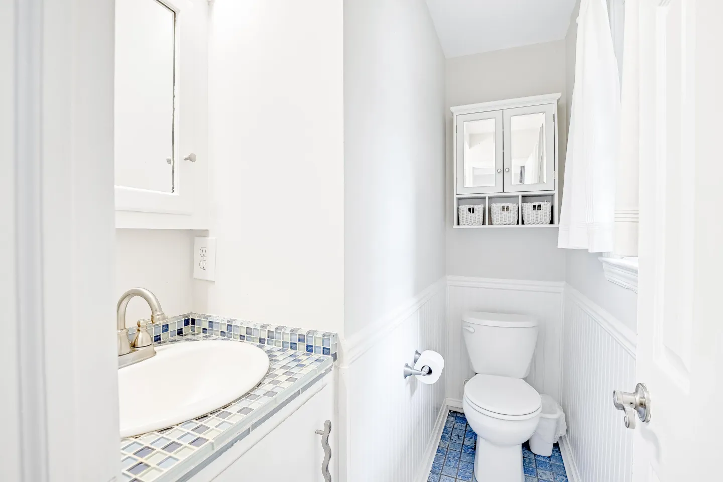 A brightly lit powder room with a tiled vanity countertop, a toilet against wainscoted walls, and a small wall-mounted cabinet.