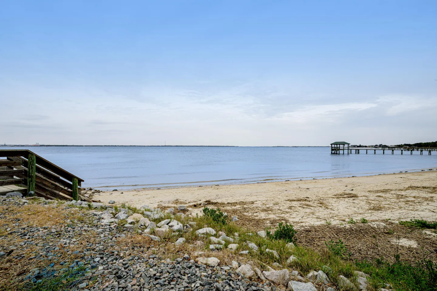 A sandy beach shoreline along calm water with wooden stairs on the left and a pier in the distance.