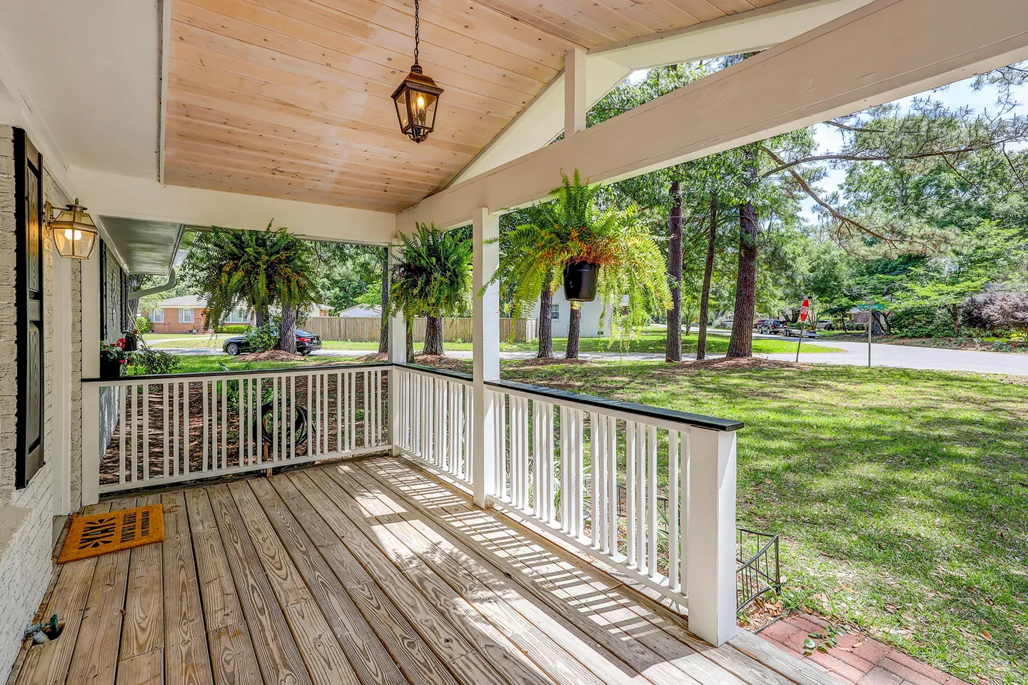 Front porch with wood-plank flooring, white railings, hanging ferns, and a view of the front yard and street.