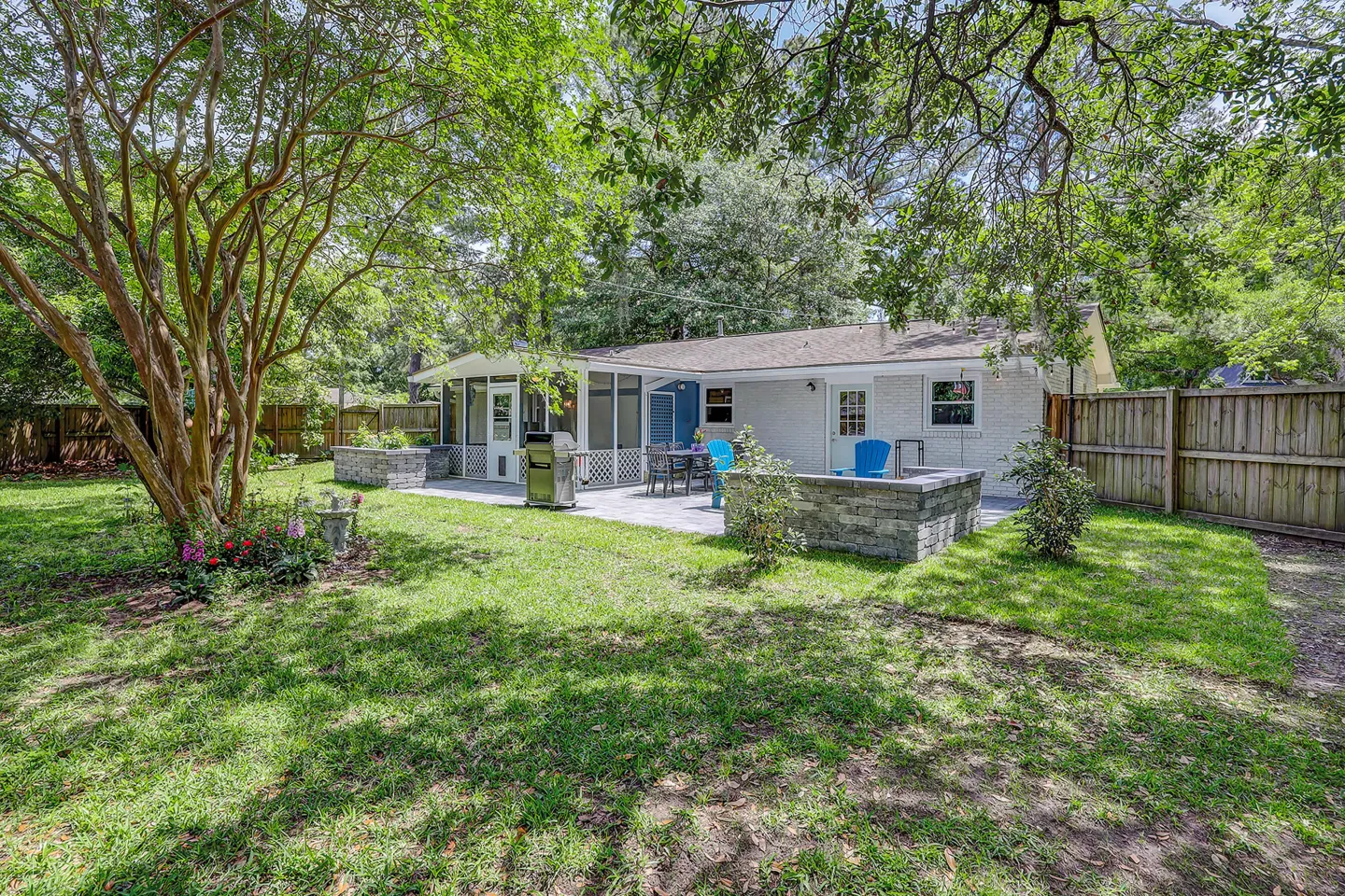 Fenced backyard featuring a paved patio with outdoor seating, a grill, low stone retaining walls, a screened porch, and mature trees.