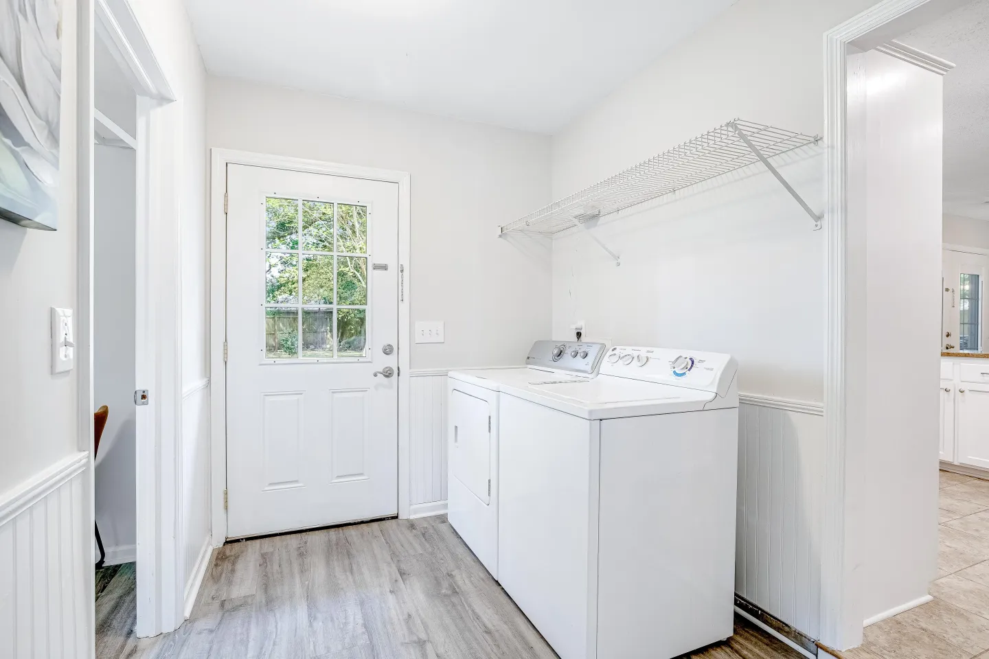 A bright laundry room featuring a side-by-side white washer and dryer, wood-look flooring, and an exterior door with window panes.