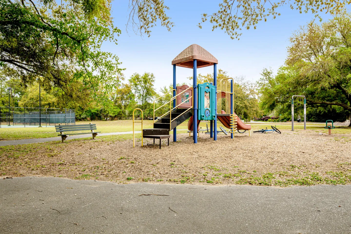 Outdoor community playground with play structure, slides, bench, and distant tennis courts.