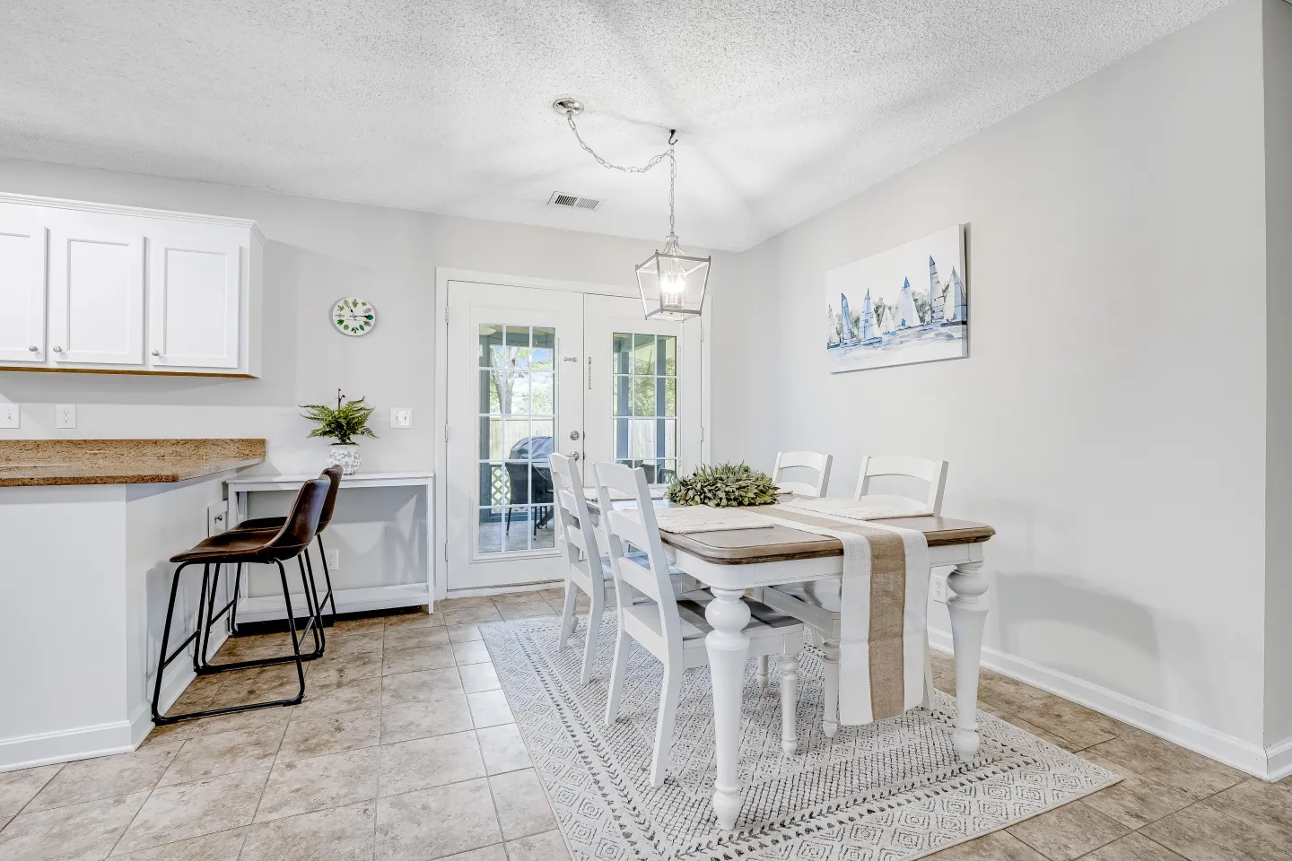 Dining area with a wooden table, white chairs, area rug, and pendant light adjacent to a kitchen counter and glass patio doors.