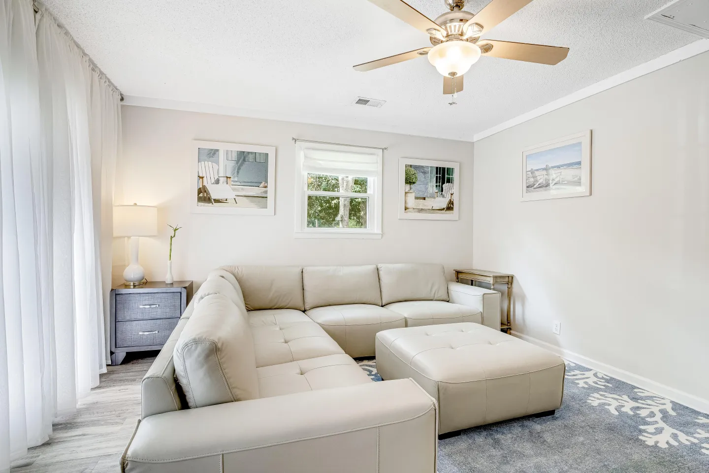 Bright living room with a cream leather sectional sofa, matching ottoman, ceiling fan, and framed coastal prints on light walls.