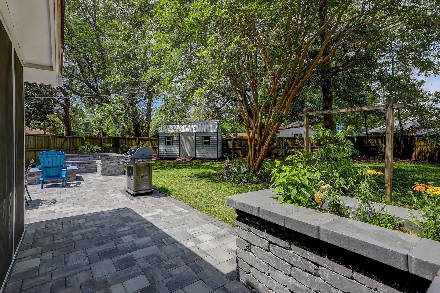 Fenced backyard featuring a paved patio with a gas grill, stone planters, fire pit area, lawn, and a storage shed under mature trees.