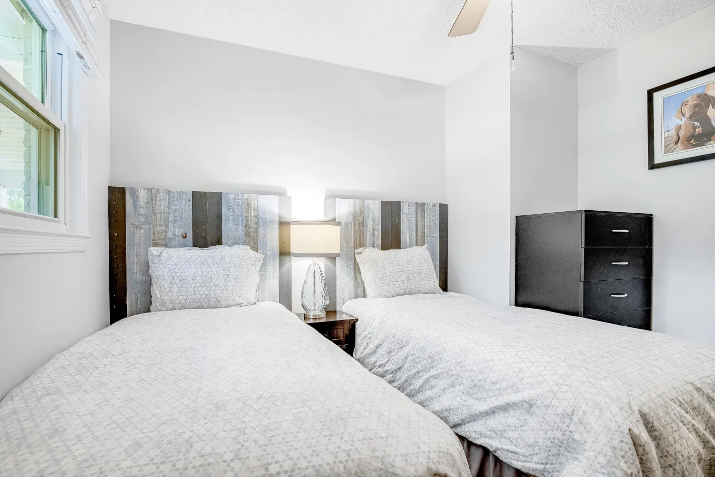 Bedroom featuring two twin beds with rustic paneled headboards separated by a nightstand and lamp, with a window on the left and a dark dresser on the right.