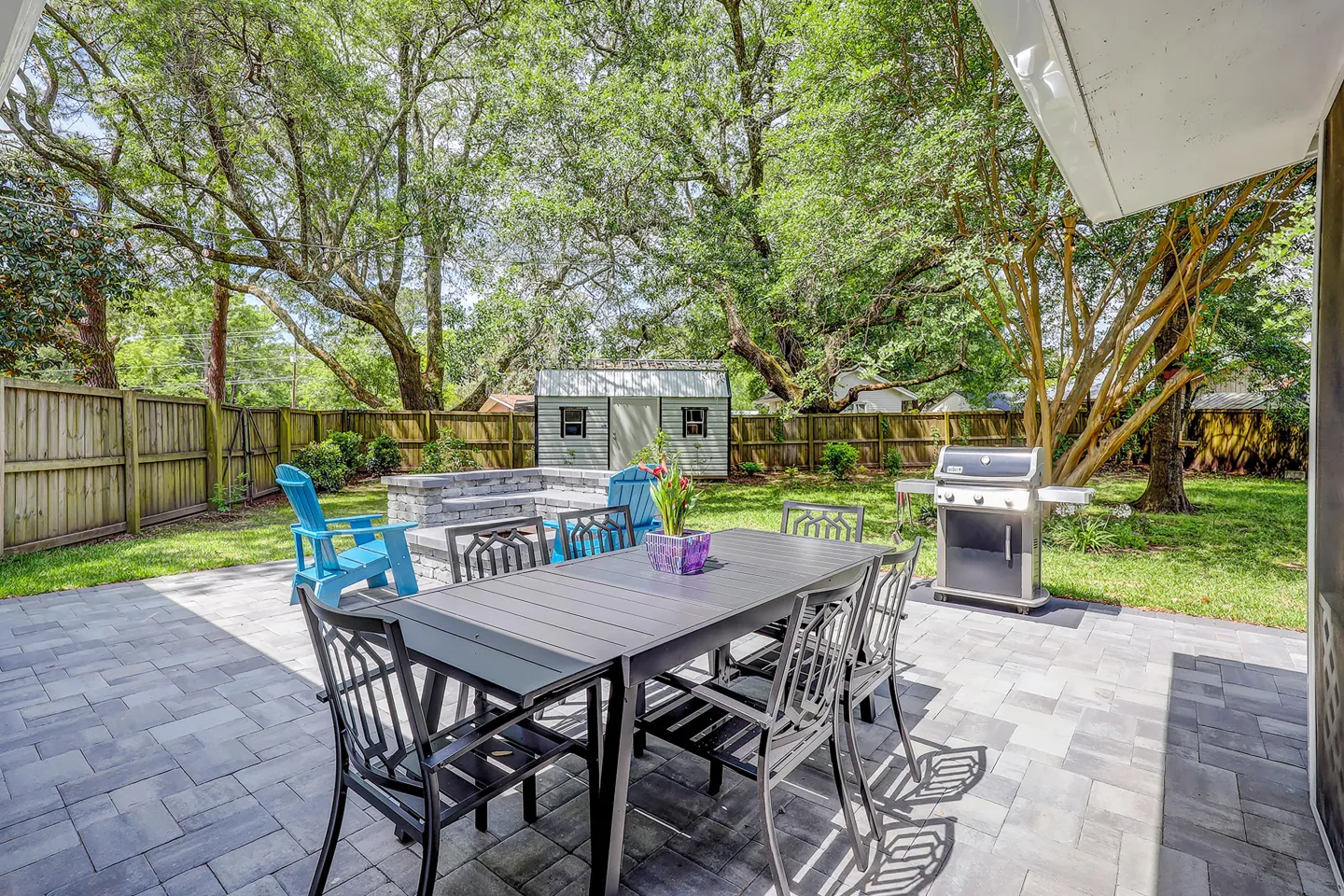 Paved backyard patio with a dining table and chairs, a barbecue grill, and a fire pit area surrounded by grass, trees, and a wooden fence.