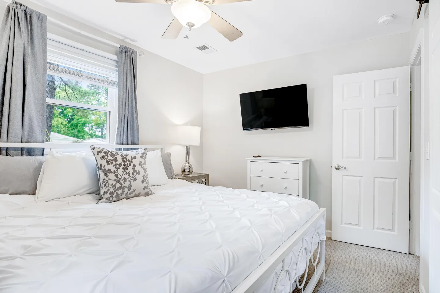 Bedroom with a white metal bed, mounted TV, small white dresser, and window with gray curtains.